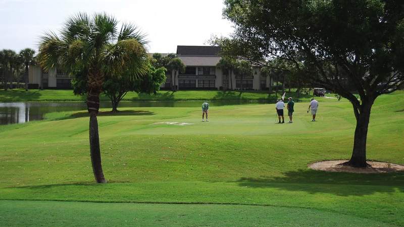 002 -Three people playing golf at Vista Plantation on a sunny day; the course features lush greens, a pond, and residential buildings in the background_