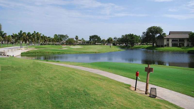 004 - A lush Vista Plantation golf course with a pond, sand bunkers, and palm trees under a clear blue sky, flanked by a pathway and residential buildings_