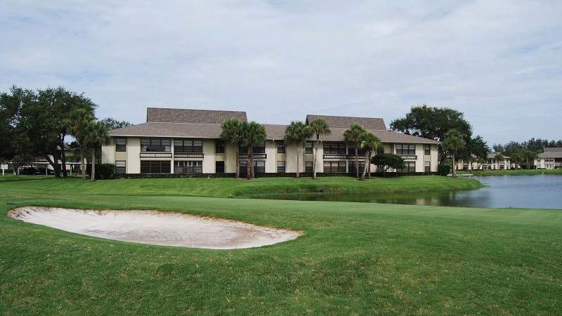 009 - Vista Plantation golf course with a sand bunker in the foreground, a calm pond in the middle, and a two-story building in the background under a cloudy sky_