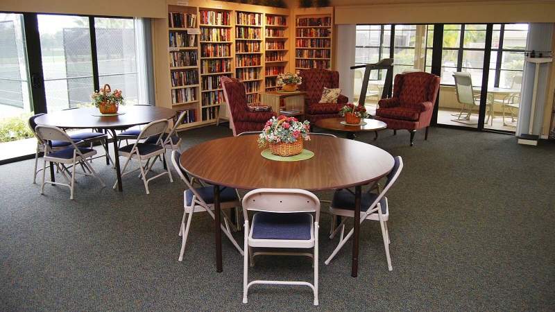 011 - Vista Plantation clubhouse featuring a cozy library lounge with multiple tables and chairs, floral centerpieces, and floor-to-ceiling bookshelves filled with books, illuminated by