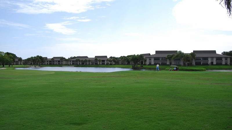 016 - A serene pond in front of low-rise Vista Plantation residential buildings with a lush green lawn and a few people in the distance under a partly cloudy sky_