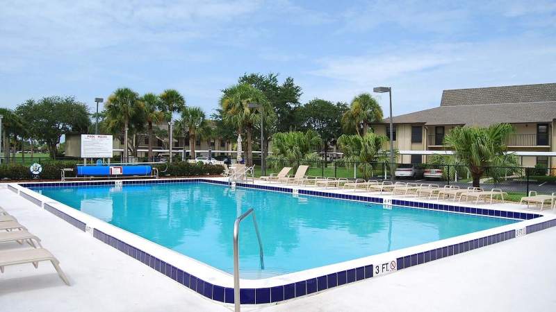 020 - A Vista Plantation outdoor swimming pool surrounded by lounge chairs and palm trees, adjacent to a residential building under a clear sky_