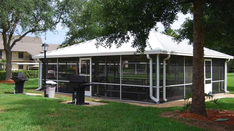 022 - A screen-enclosed outdoor patio area with tables and chairs, surrounded by grass and trees, near residential buildings_