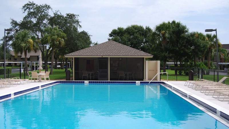 027 - A Vista Plantation community pool with a blue water surrounded by a white fence, adjacent to a small building with chairs under a roof, set in a grassy area with trees in the back