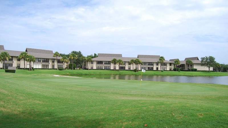 029 - Vista Plantation golf course with a large green in the foreground, a flag at the hole, and a line of two-story buildings beside a lake under a bright blue sky_