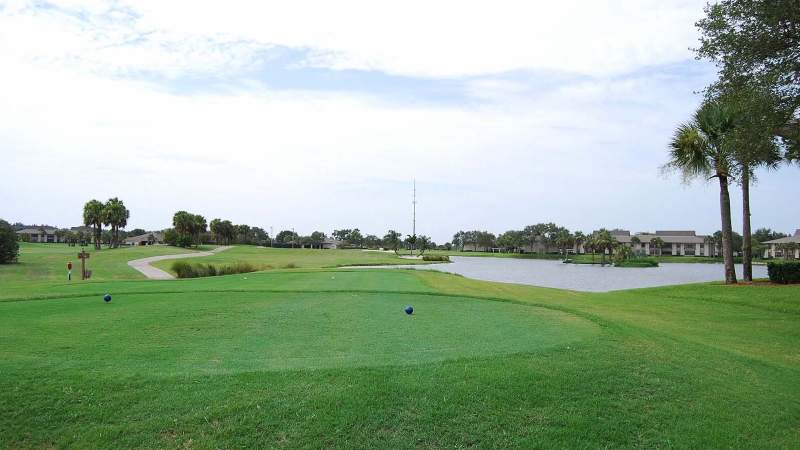 030 - Vista Plantation golf course with vibrant green grass, a water hazard on the right, and suburban buildings in the background under a cloudy sky_